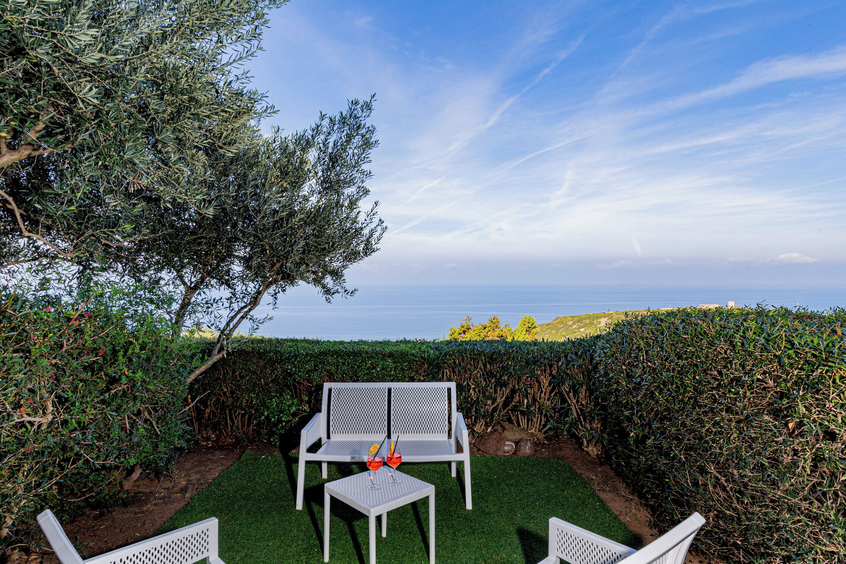 An outdoor seating area with white chairs and a table, surrounded by greenery and a view of the sea.