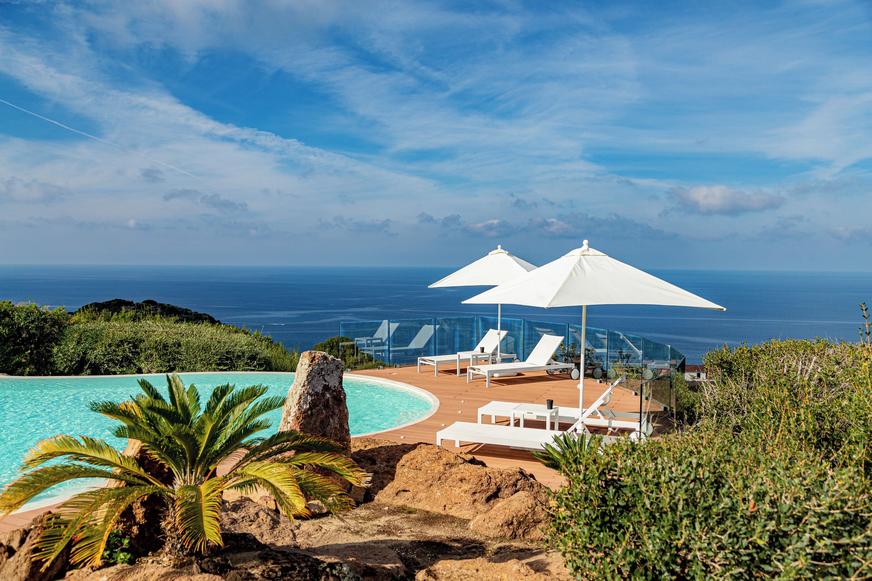 Zona de piscina con sillones reclinables y sombrillas, con vista a un océano azul y despejado.