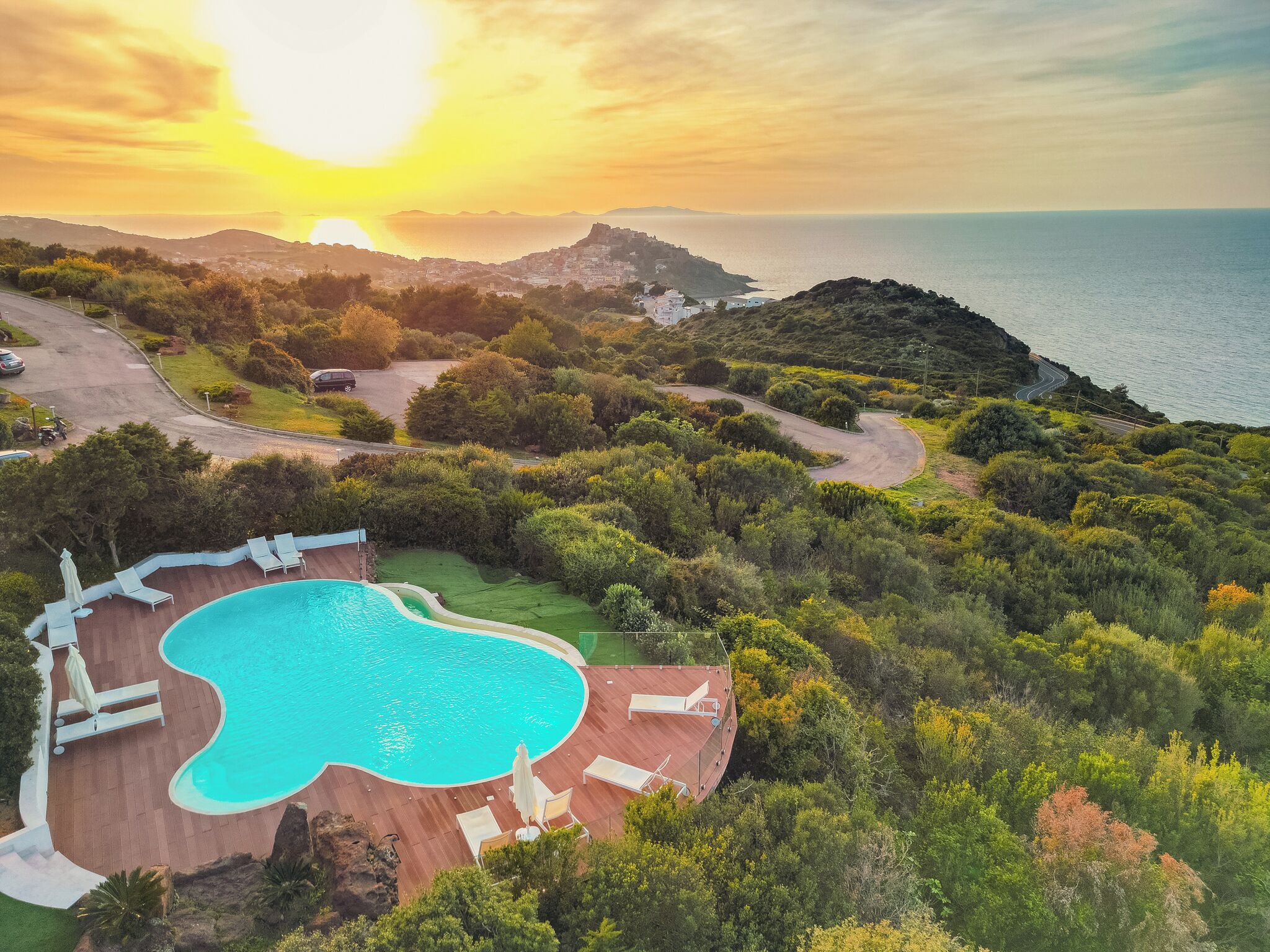 Una piscina rodeada de exuberante vegetación, un camino serpenteante y una vista al mar al atardecer.