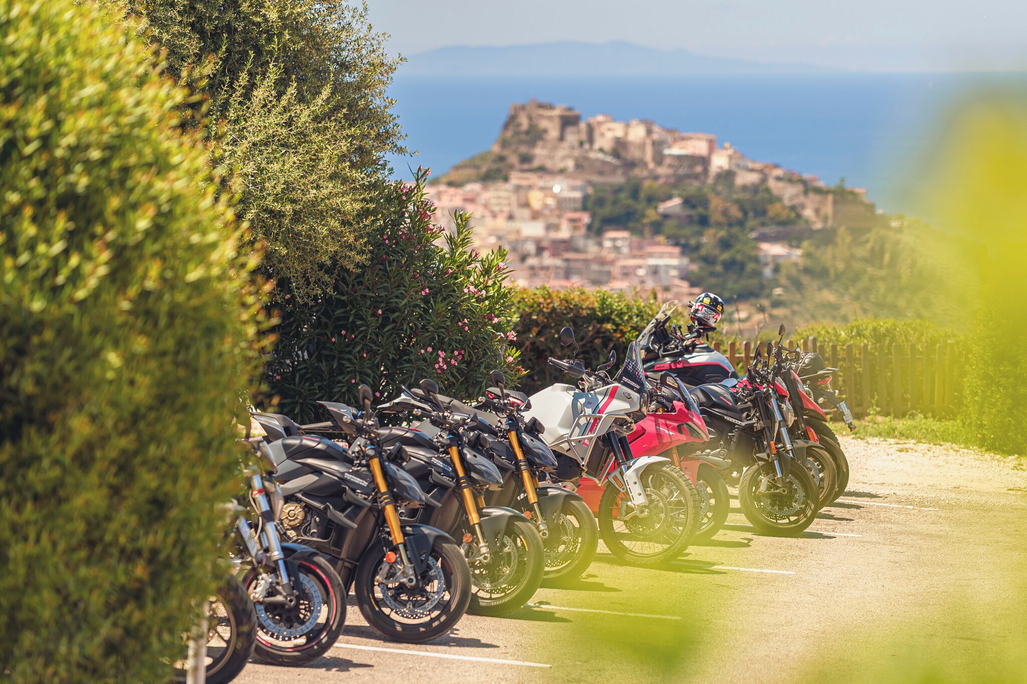 Uma fileira de motocicletas estacionadas em uma estrada, com vista para um assentamento histórico no alto de uma colina.