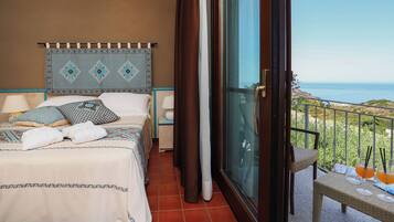 A bedroom with a bed, pillows, a nightstand, a window with curtains, and a balcony with a view of the sea.
