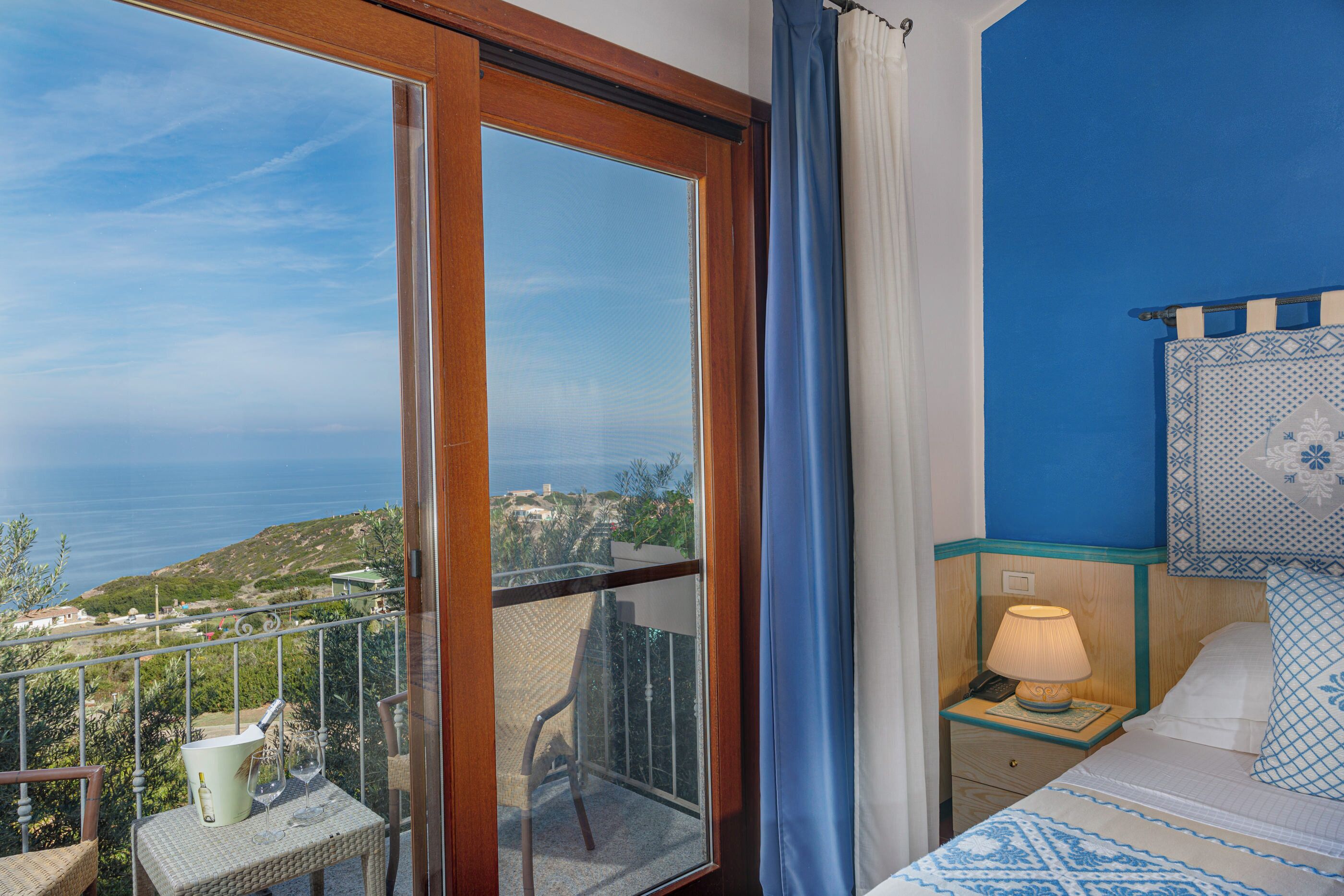 Un balcone con vista sul mare, un letto con biancheria a motivi decorativi e un comodino con una lampada.