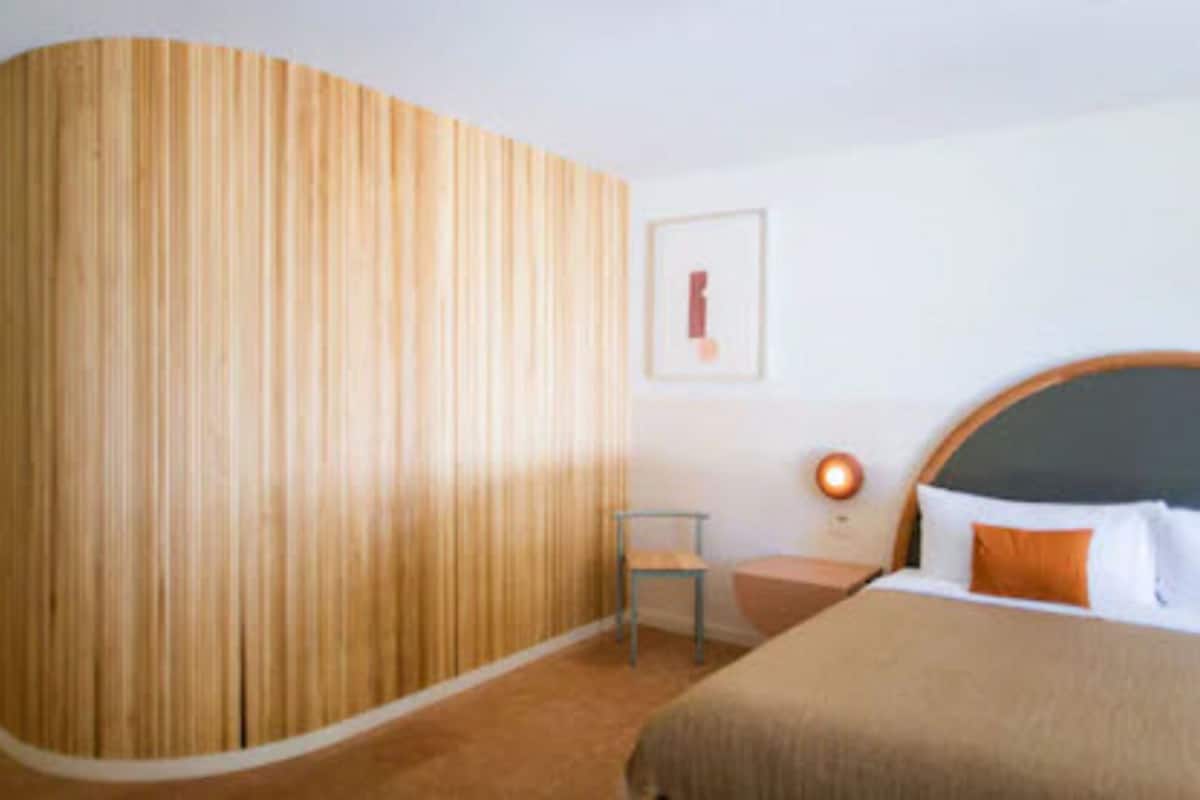 A hotel room with a curved wooden wall, a bed with an orange pillow, a small chair, and a bedside table with a lamp.