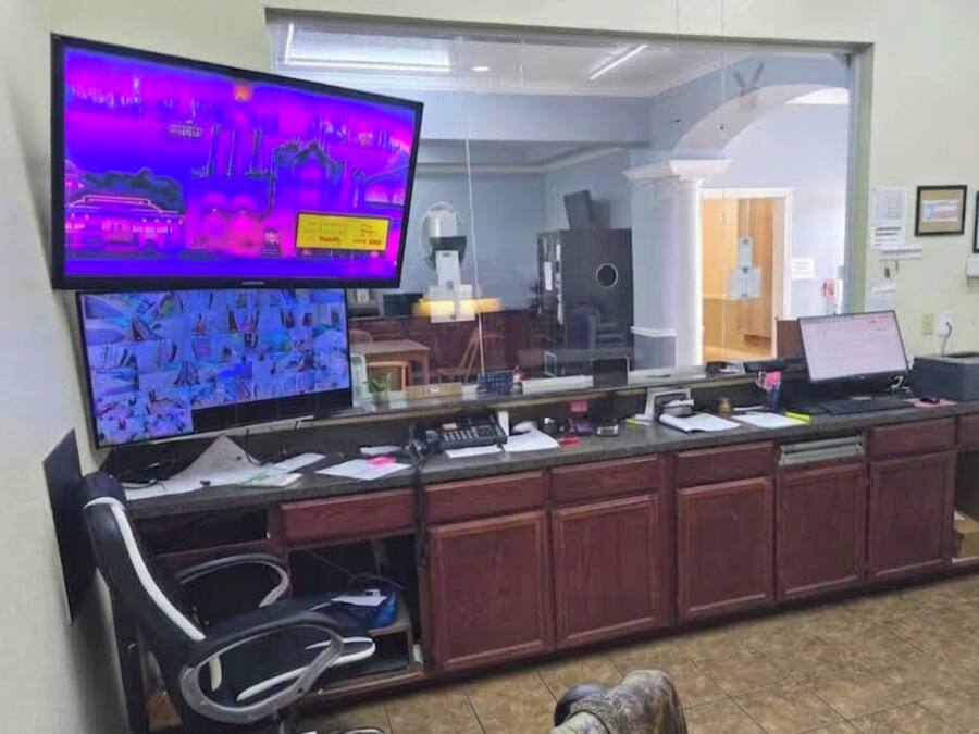 A reception area with a large flat-screen TV mounted on the wall, a reception desk with a computer and paperwork, a chair, and a mirror reflecting the interior.