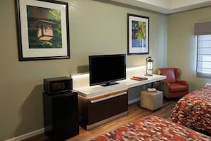 A hotel room with a flat-screen TV, two beds, a small cabinet, a chair, and framed pictures on the wall.