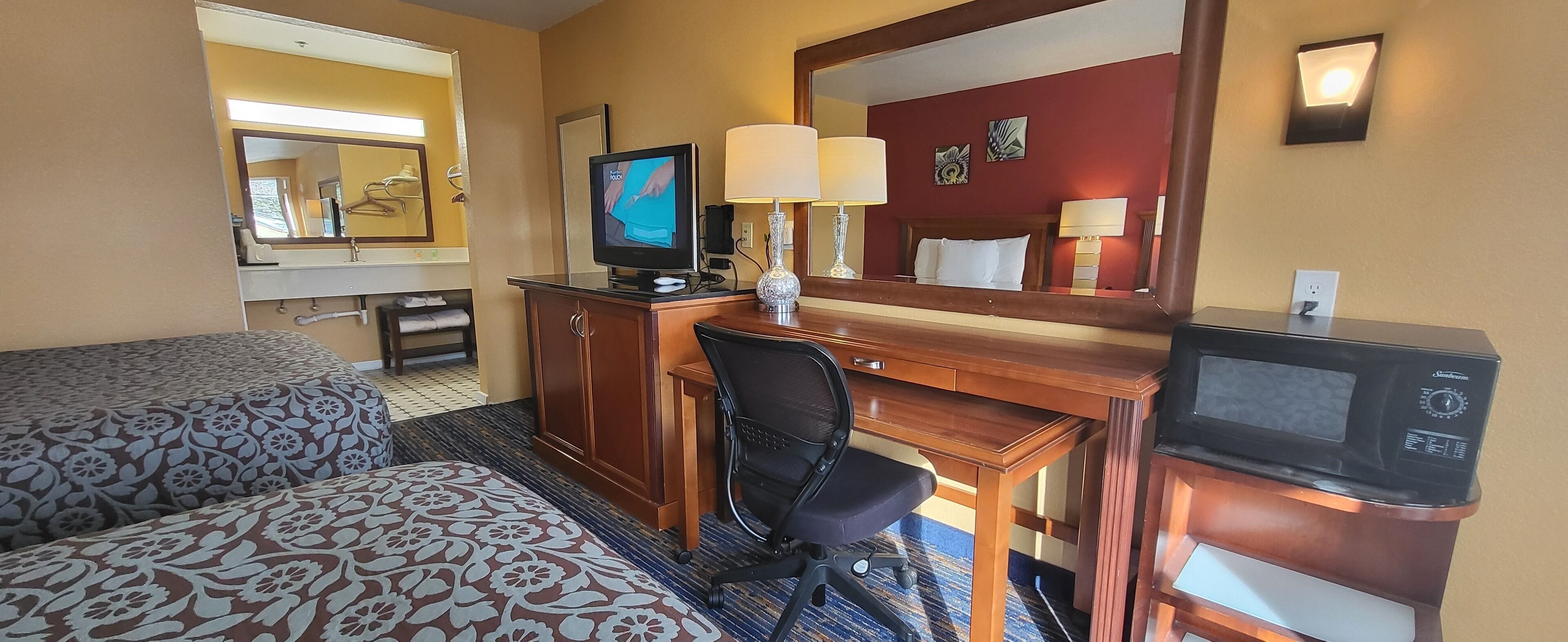 A hotel room with a bed, a desk with a computer and a television, a chair, a microwave, and a mirror.