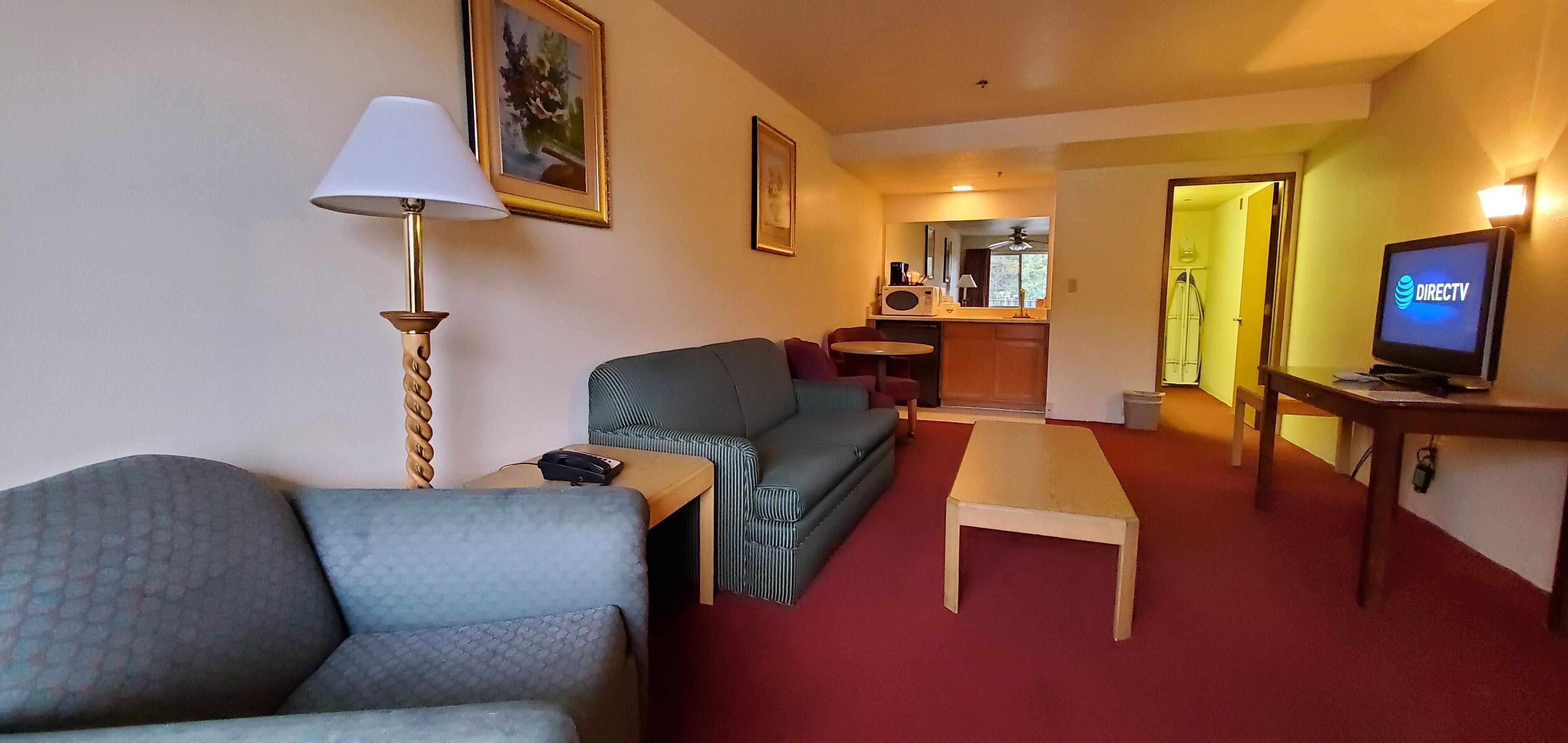 A hotel room with a sofa, a small table, a lamp, a television, and a kitchen area in the background.