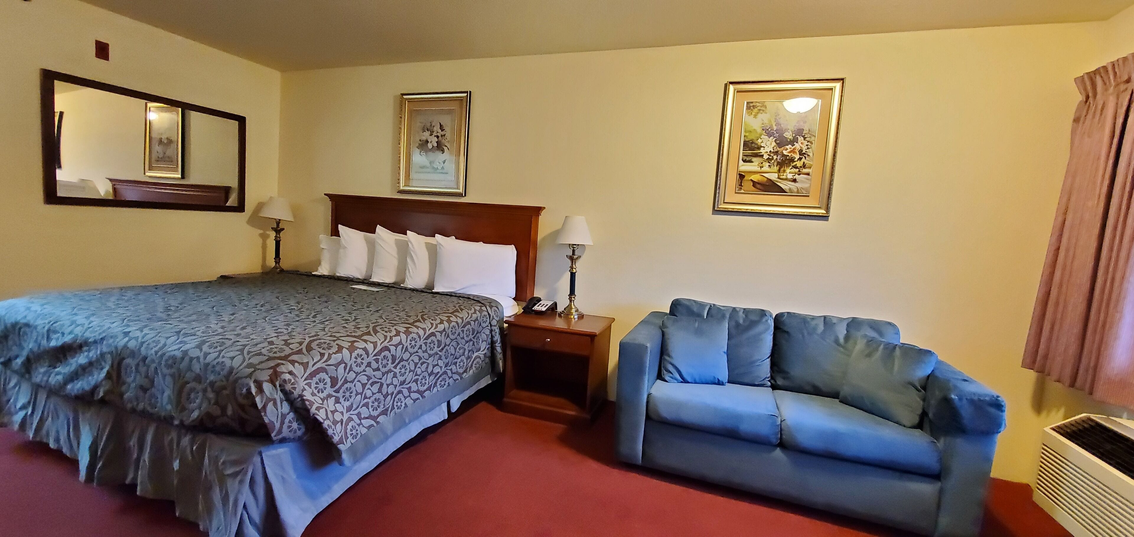 A hotel room with a bed, a blue sofa, a nightstand with a lamp, and two framed pictures on the wall.