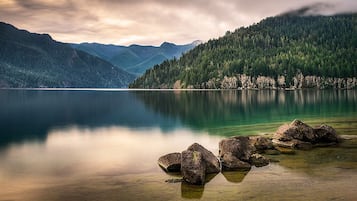 A calm lake with clear water, surrounded by forested mountains and rocky outcrops.