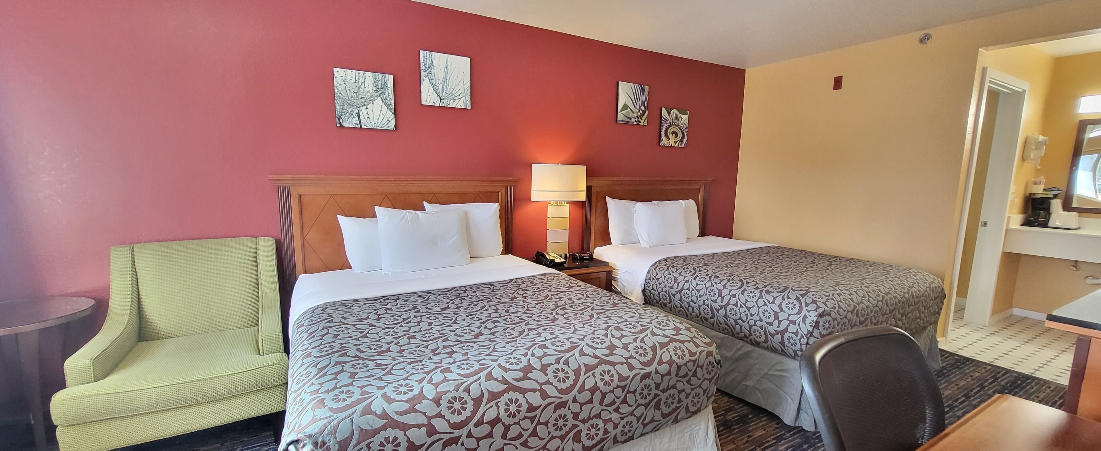 A hotel room with two beds, a green armchair, a bedside table with a lamp, and artwork on the walls.