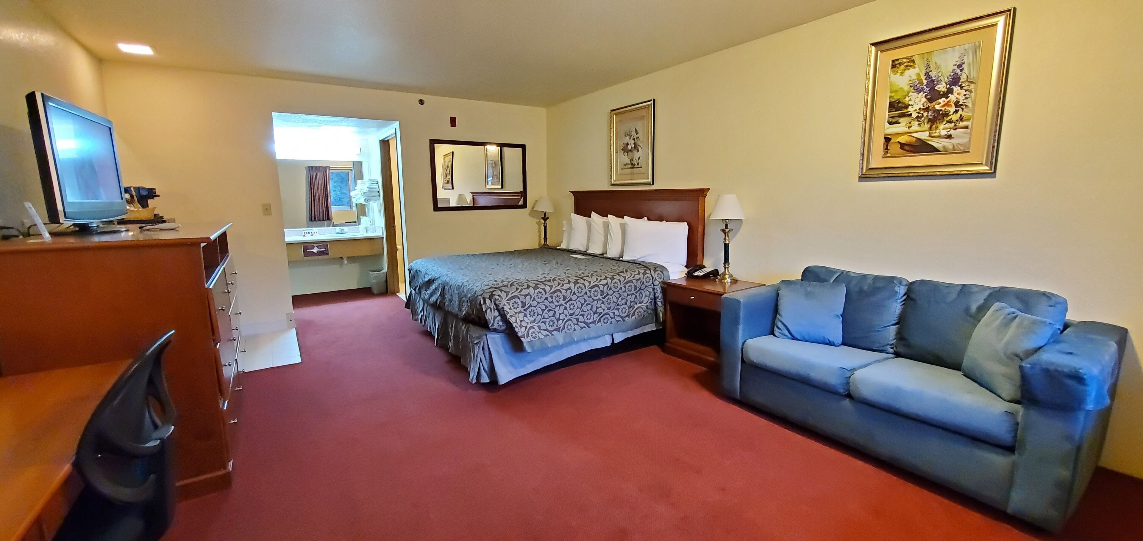 A hotel room with a bed, a blue sofa, a desk with a computer, a mirror, and a painting on the wall.