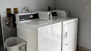 A laundry area with two front-loading washing machines, a trash bin, and a wall-mounted electrical outlet. - General Bragg Inn & Suites Chickamauga (Chickamauga)