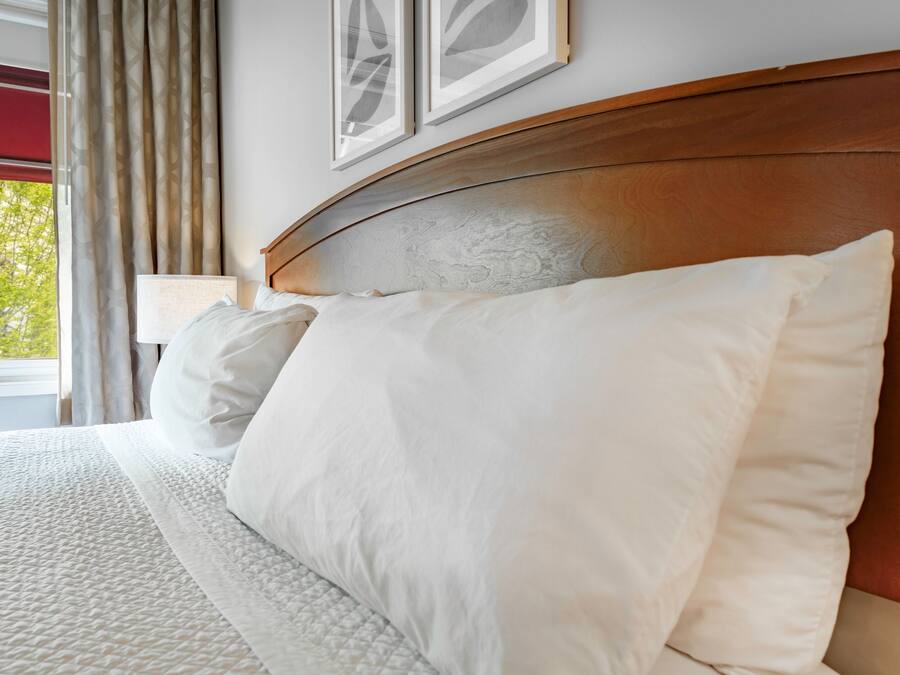 A neatly made bed with white bedding and a wooden headboard, a window with curtains, and framed artwork on the wall.