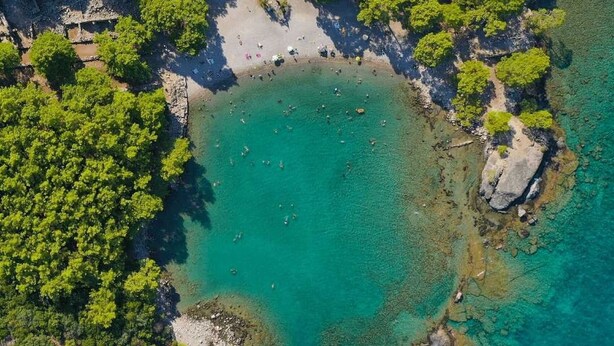 An aerial view of a beach with clear turquoise water, surrounded by dense green foliage and rocky outcrops. - Hotel Lunay (Konyaalti)