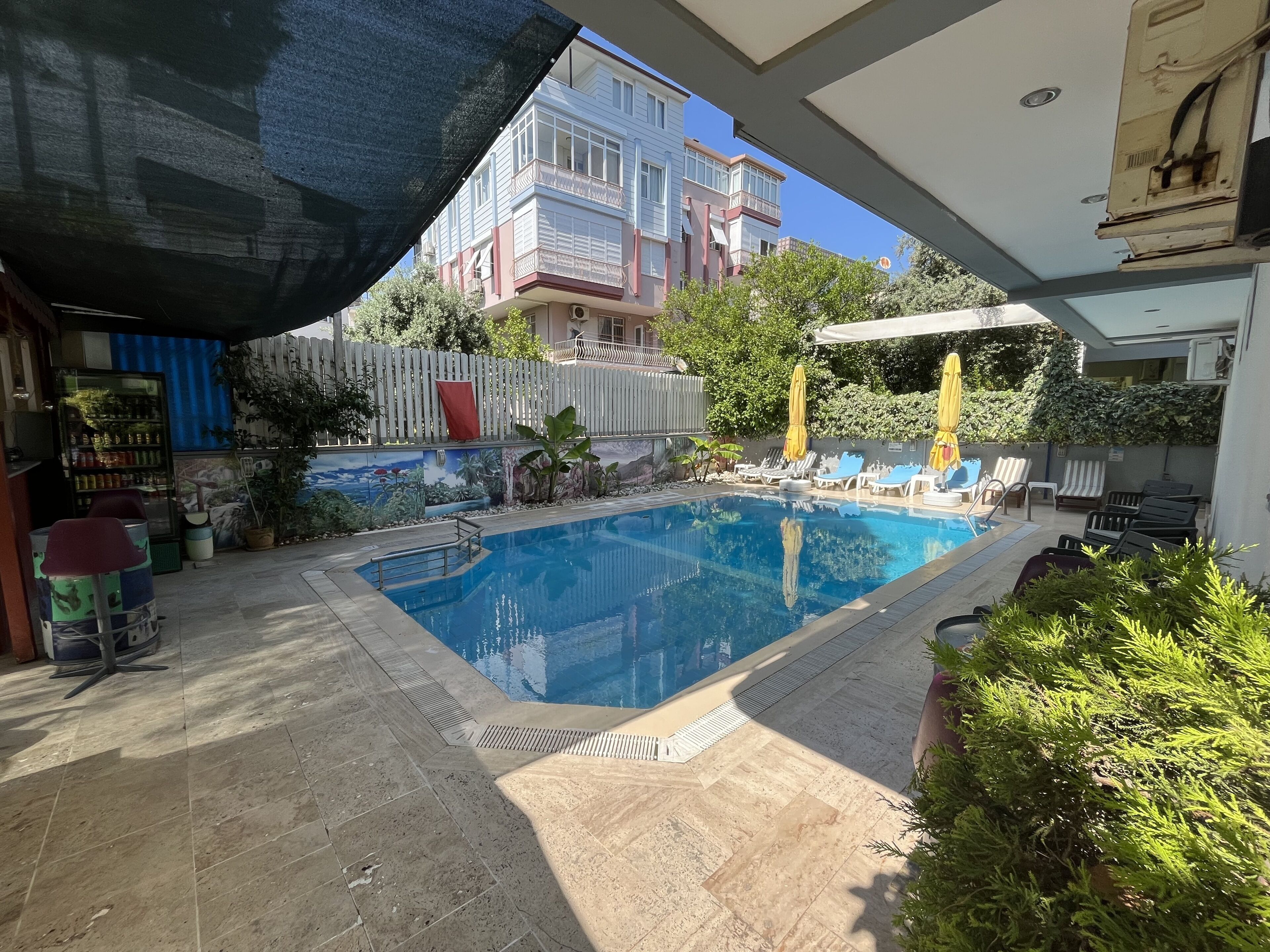 Una piscina con sillas de descanso y sombrillas, rodeada por un patio con baldosas y una pared con murales.