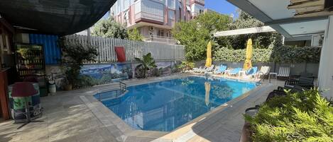 Una piscina con sillas de descanso y sombrillas, rodeada por un patio con baldosas y una pared con murales.