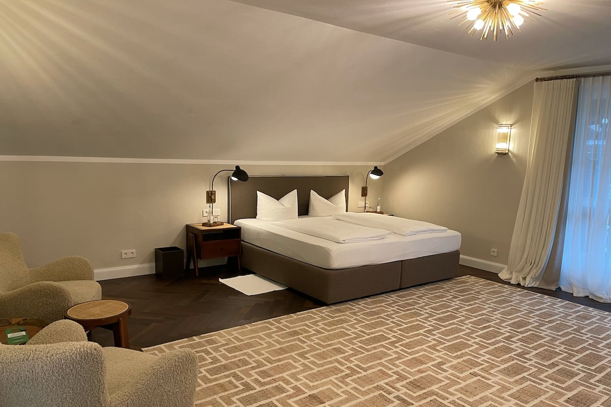 A bedroom with a large bed, a nightstand with a lamp, a chair, and a patterned rug.