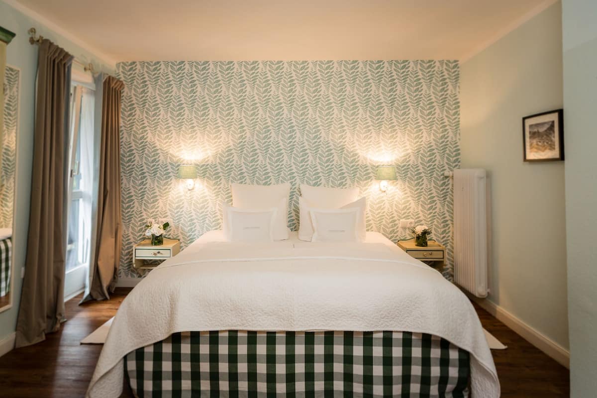 A neatly made bed with a checkered bed skirt, two bedside tables with lamps, and a patterned wallpaper.