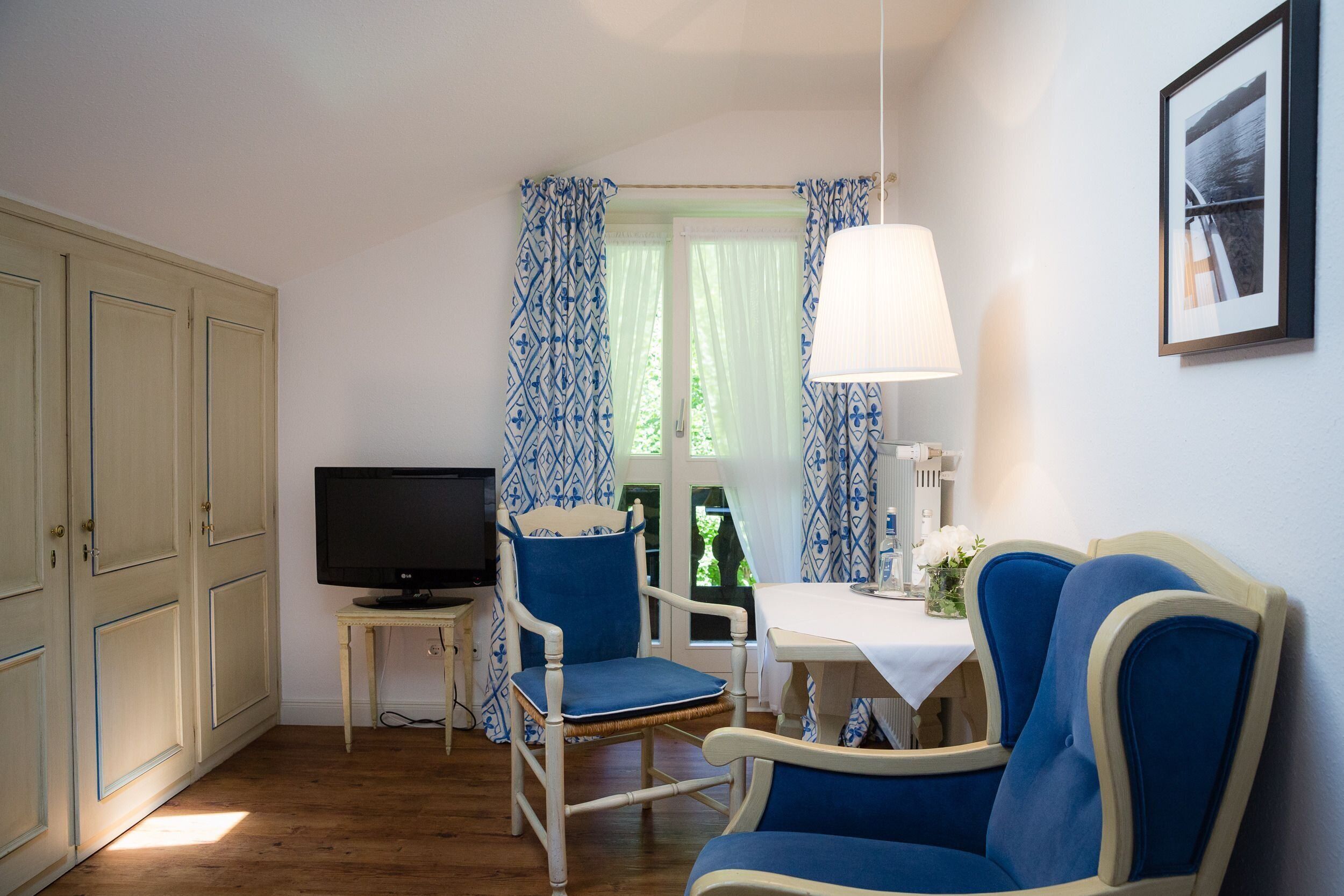 Une pièce avec une télévision, deux chaises bleues, une petite table recouverte d’une nappe blanche et une lampe.