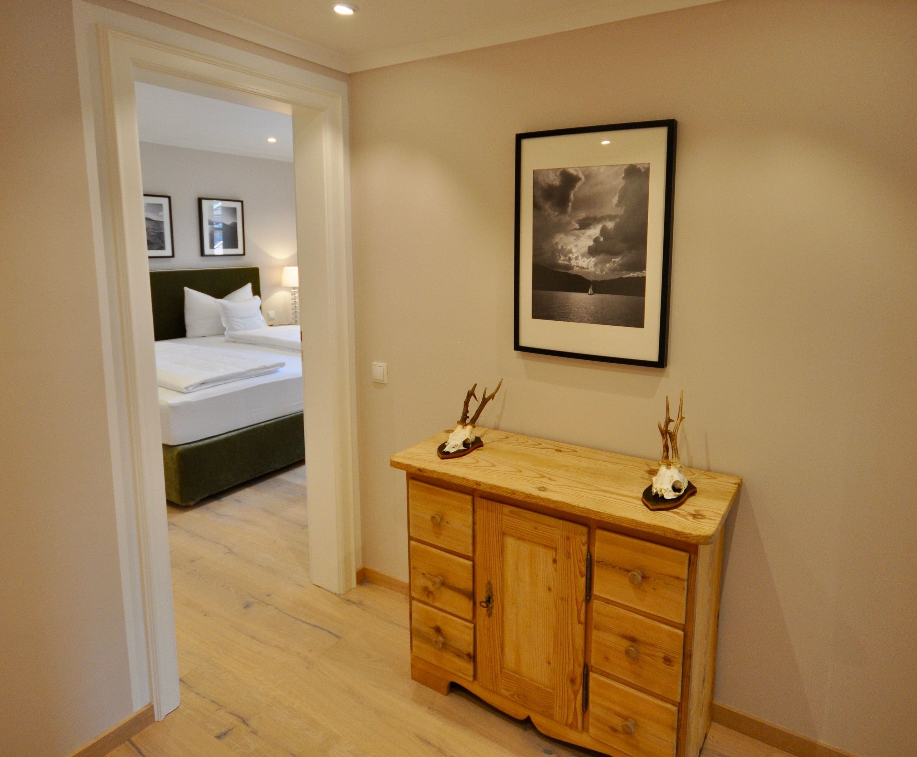 A hotel room with a bed, a wooden dresser, a framed picture on the wall, and a hallway leading to another room.