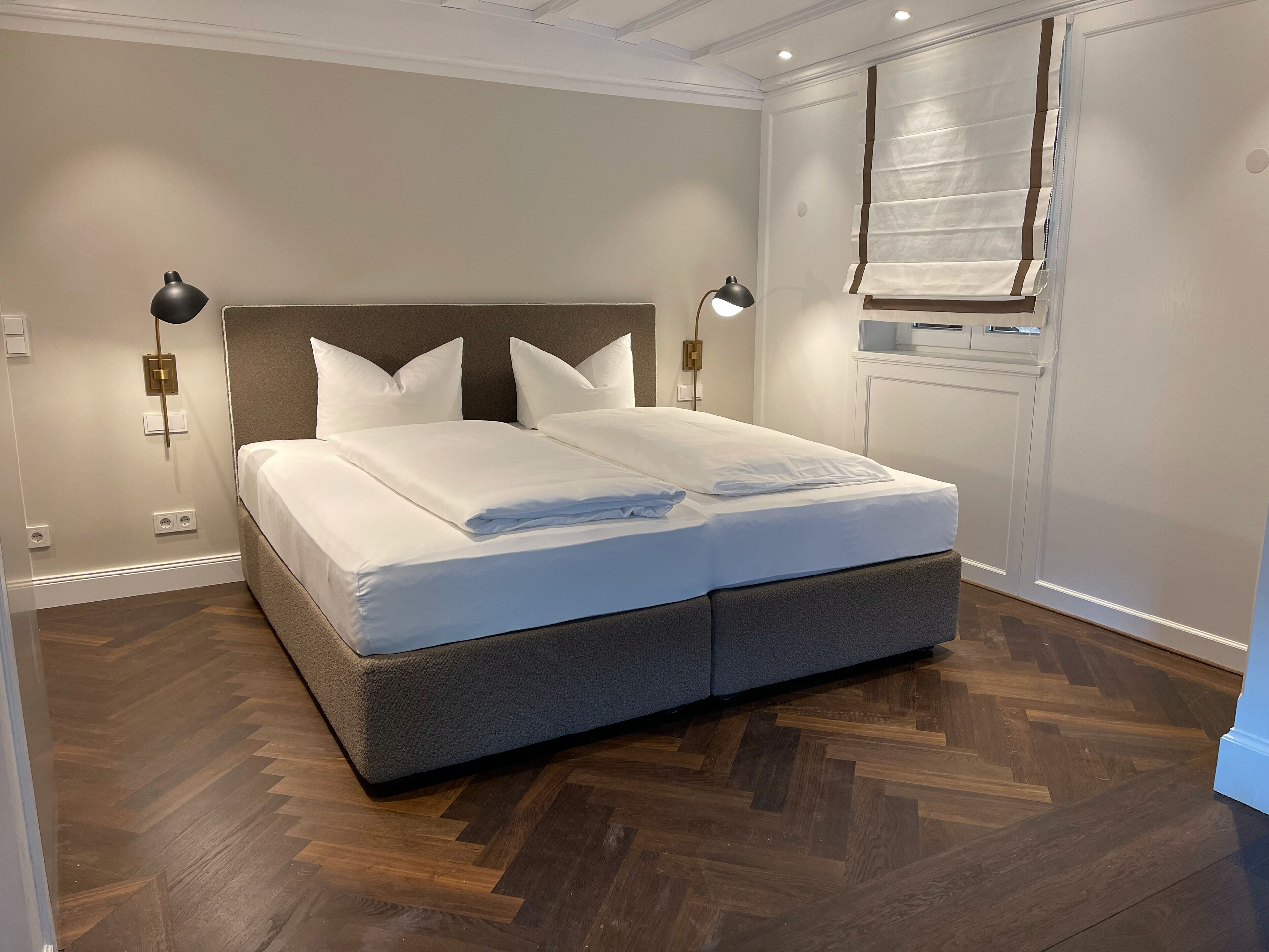 A neatly made bed with a headboard, two bedside lamps, and a window with blinds.