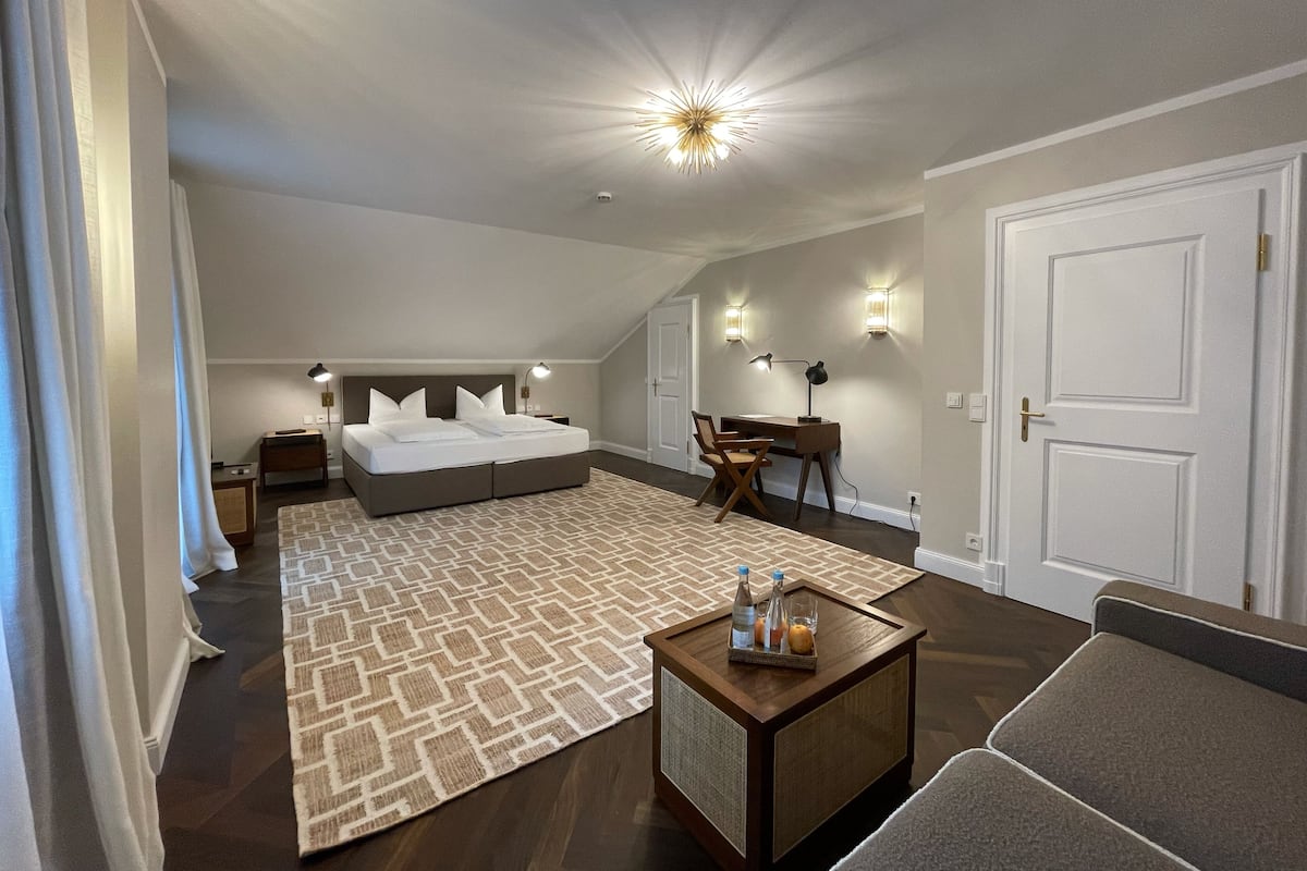 A modern hotel room with a large bed, a desk, and a chair. There is a small table with bottles and glasses on it.