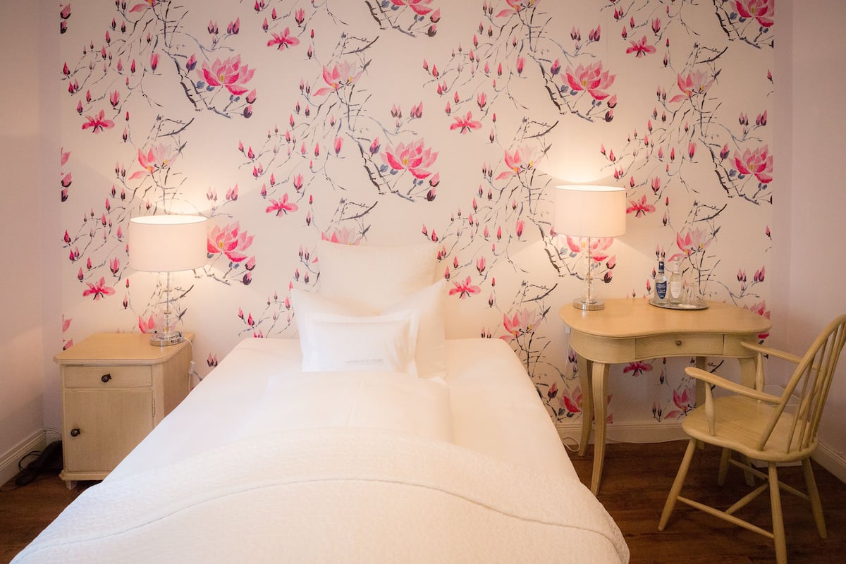 A bedroom with a bed, two bedside lamps, a wooden nightstand, a wooden desk, and a wooden chair.