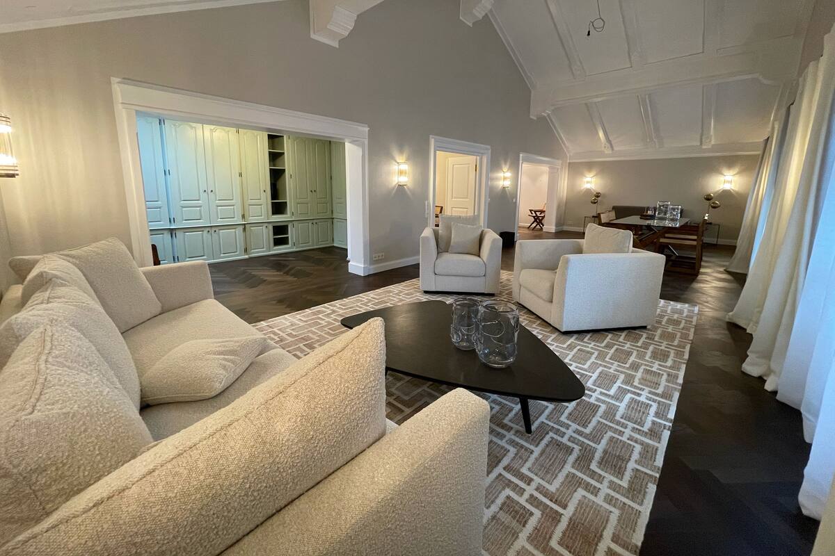 A spacious living room with a large sectional sofa, a coffee table, and a patterned rug. There is a dining area with a table and chairs, and a kitchen visible in the background.