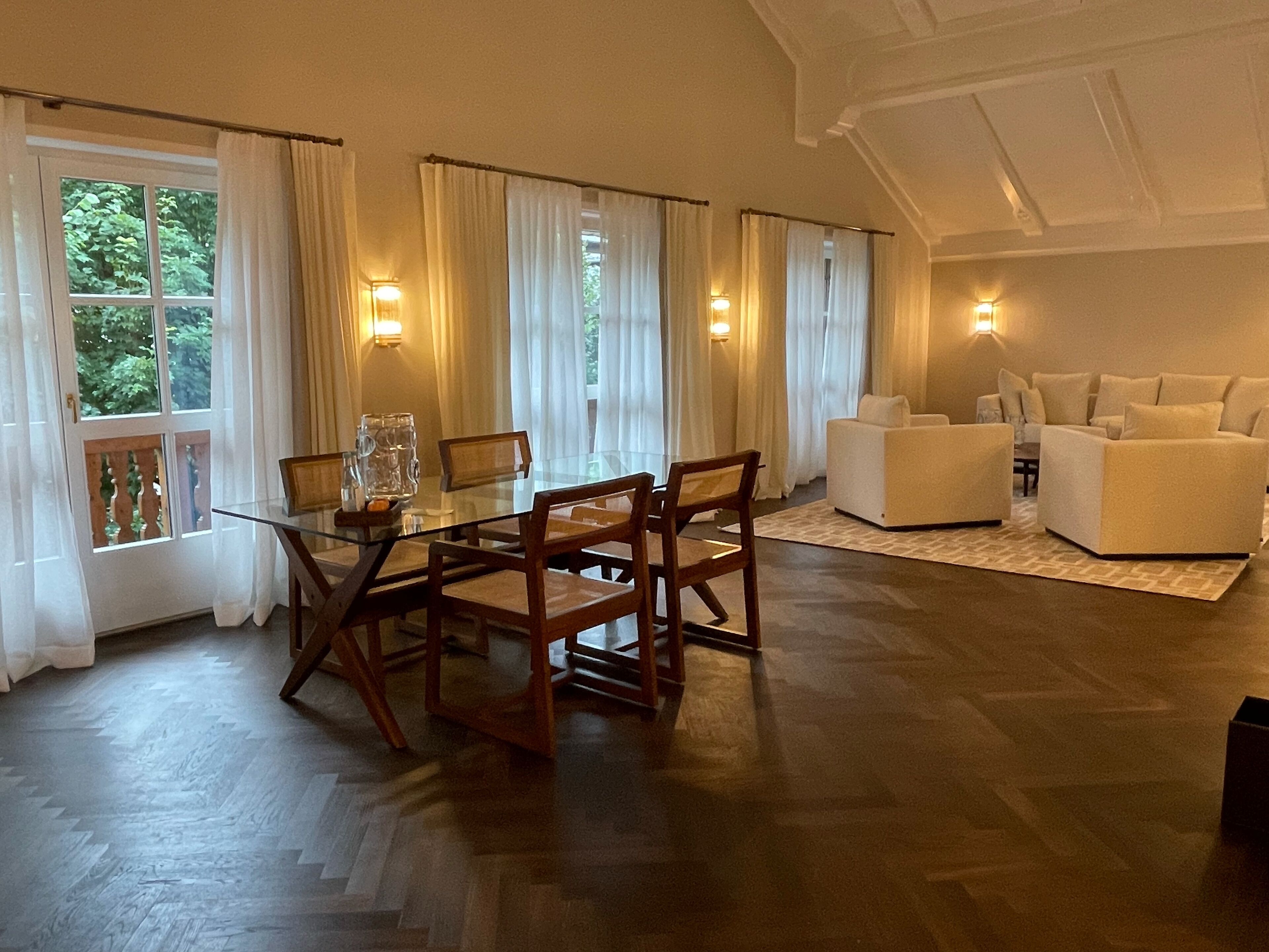 Ein Essbereich mit einem Glastisch, Holzstühlen, einer Couch und Blick auf Grün hinter großen Fenstern.