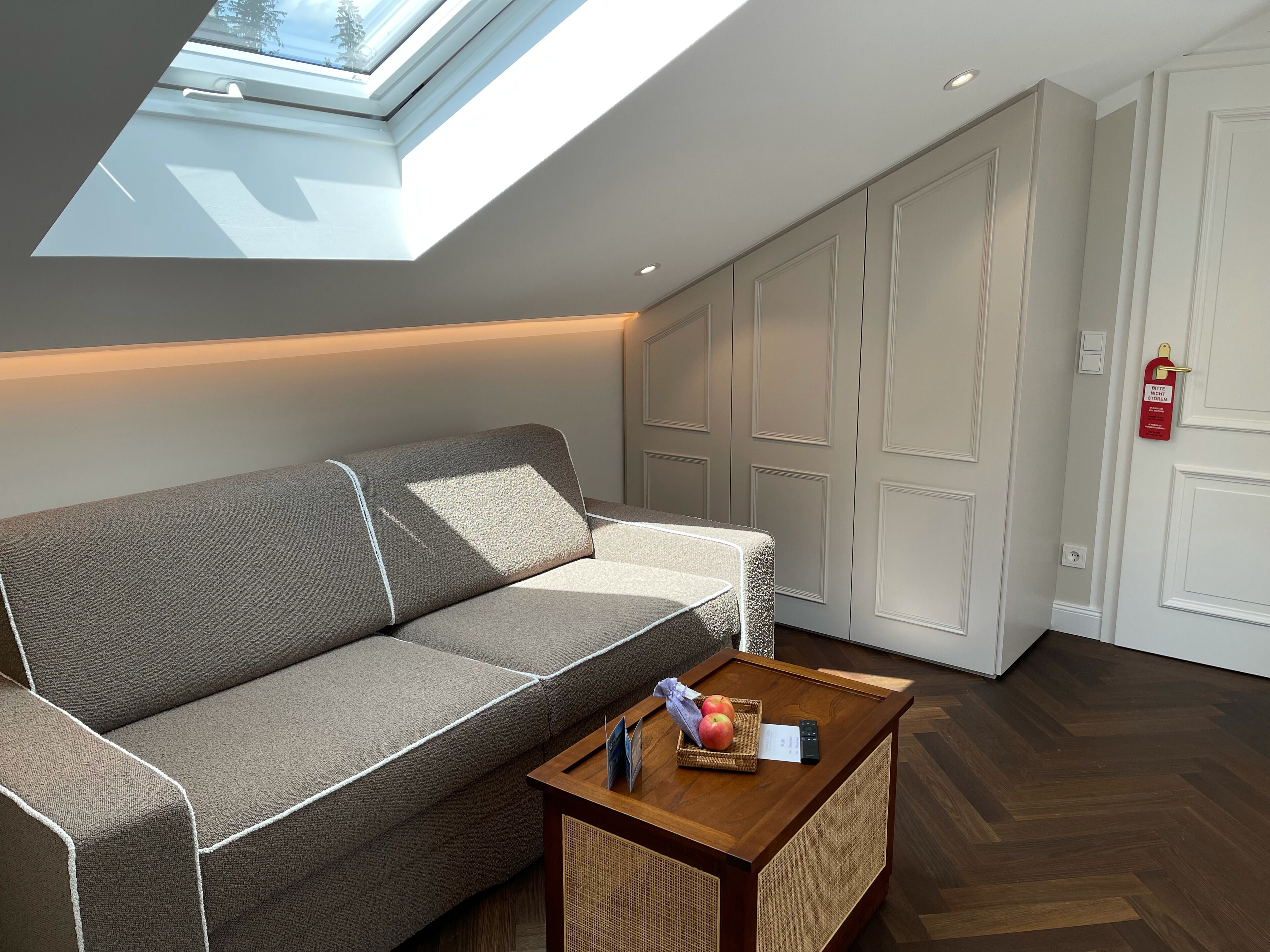 Een moderne woonkamer met een grijze bank, een houten salontafel met appels en een afstandsbediening, en een dakvenster erboven.