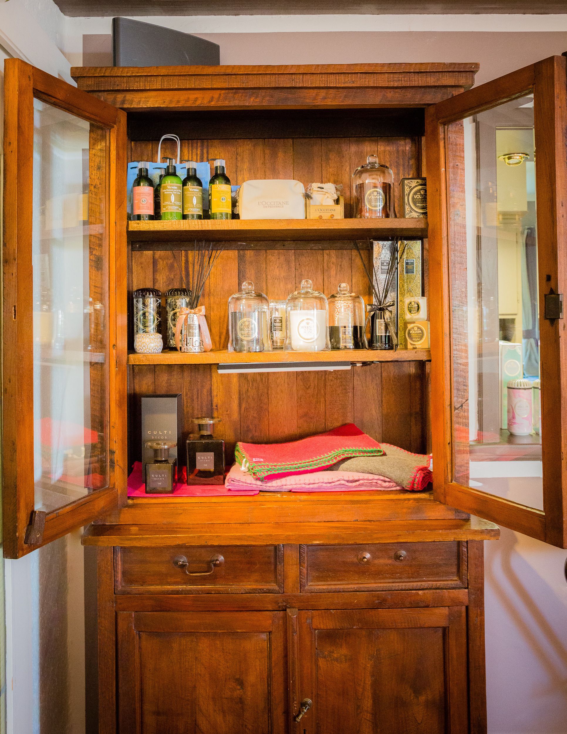 A wooden cabinet with glass doors, containing various bottles and containers, likely for toiletries or personal care products.