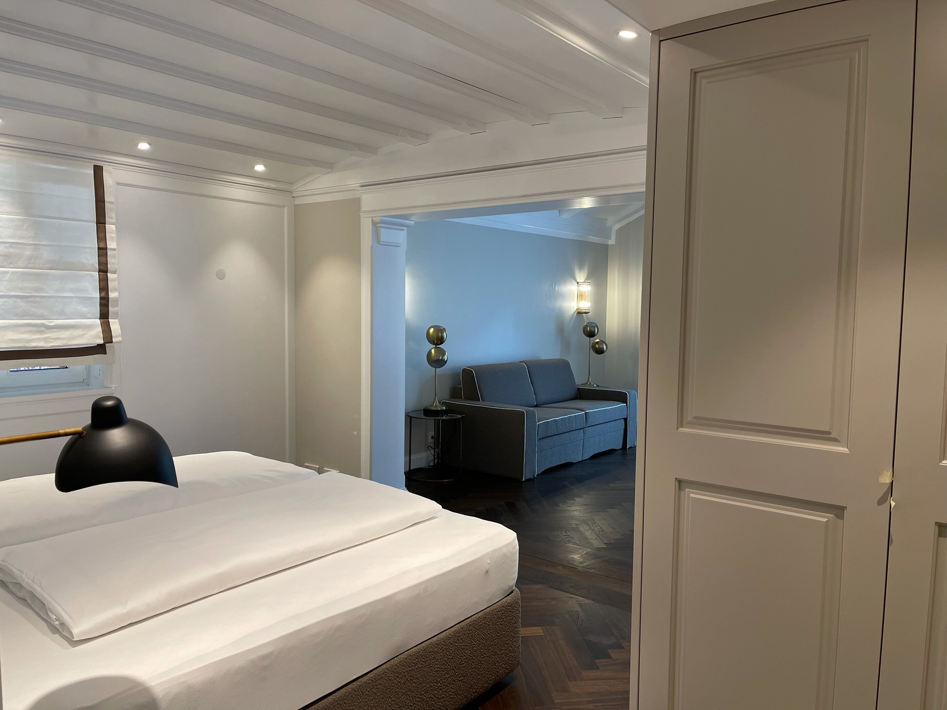 Una camera d'albergo con un letto rifatto, una lampada da comodino, un comodino e un divano nella stanza adiacente.