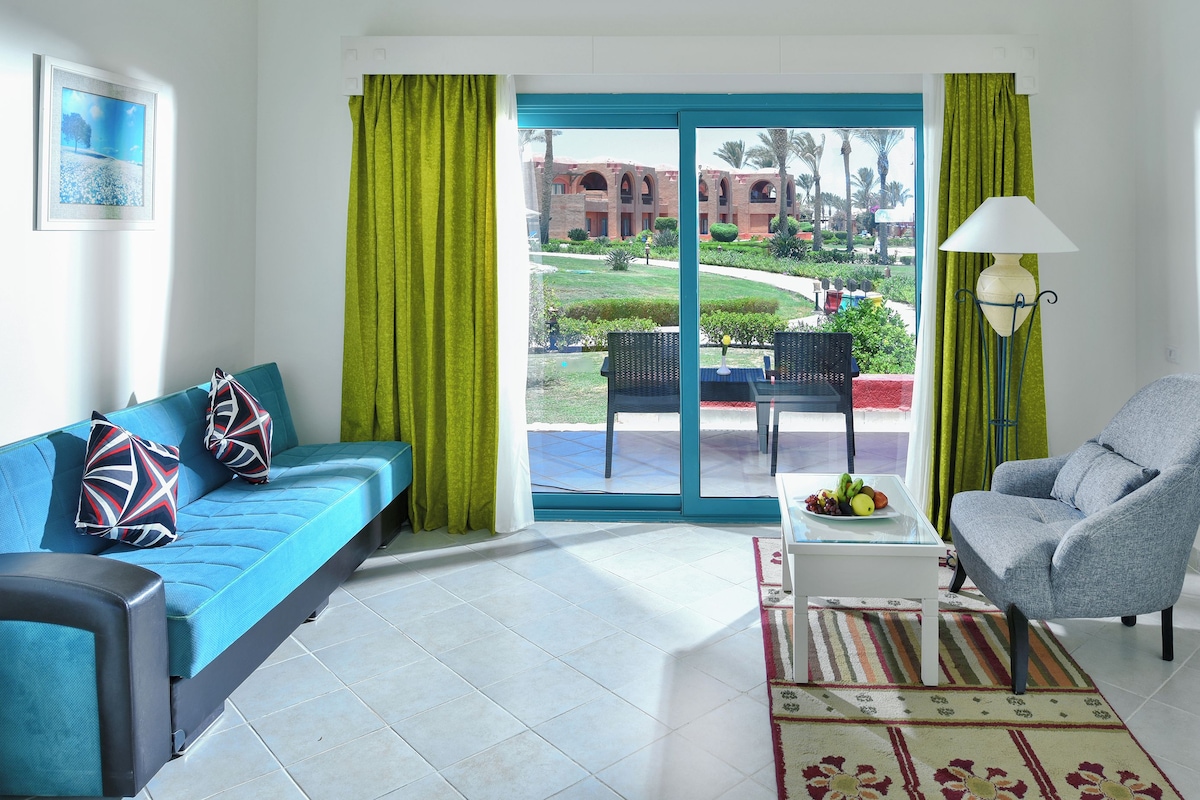 Habitación con un sofá azul, un sillón gris, una mesa de vidrio con fruta y vistas a un edificio con arcos.