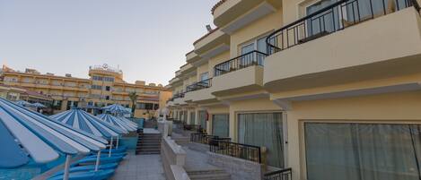 A hotel with multiple balconies, a pool area with umbrellas, and a clear sky.