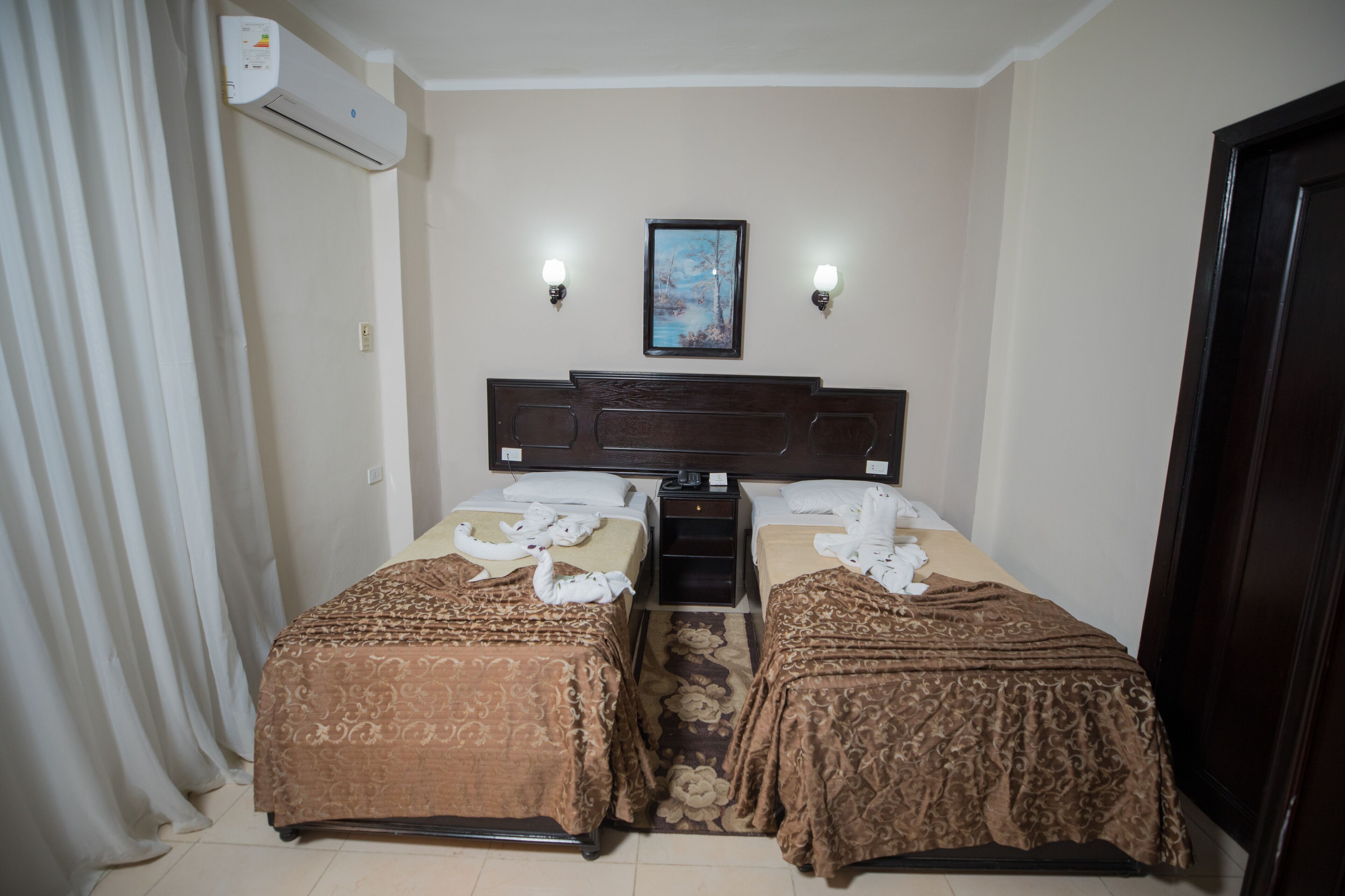 A hotel room with two beds, a large window, an air conditioning unit, and a painting on the wall.