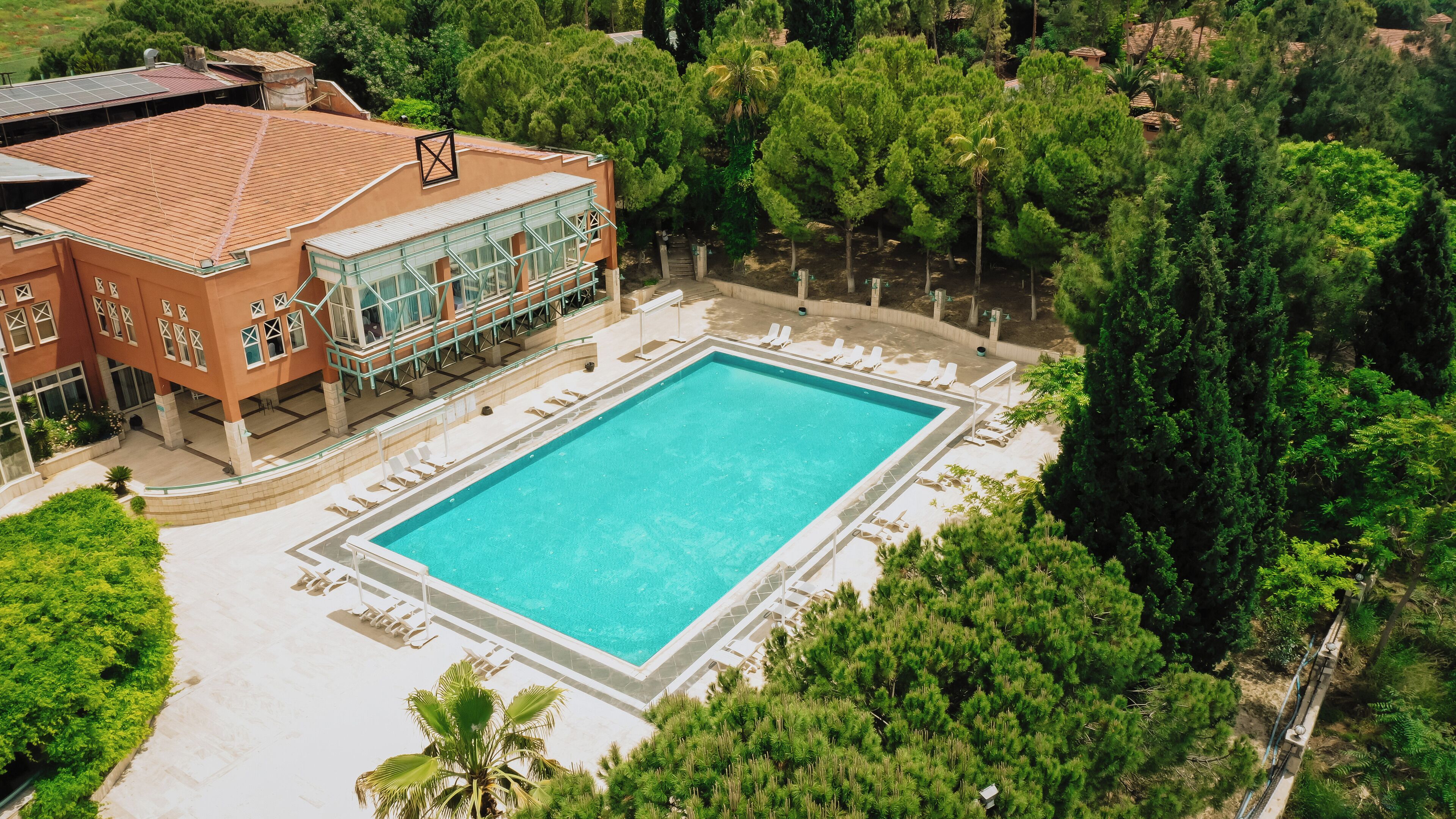 Vista aérea de una piscina rodeada de exuberante vegetación, con un edificio de grandes ventanales y techo de tejas al fondo.