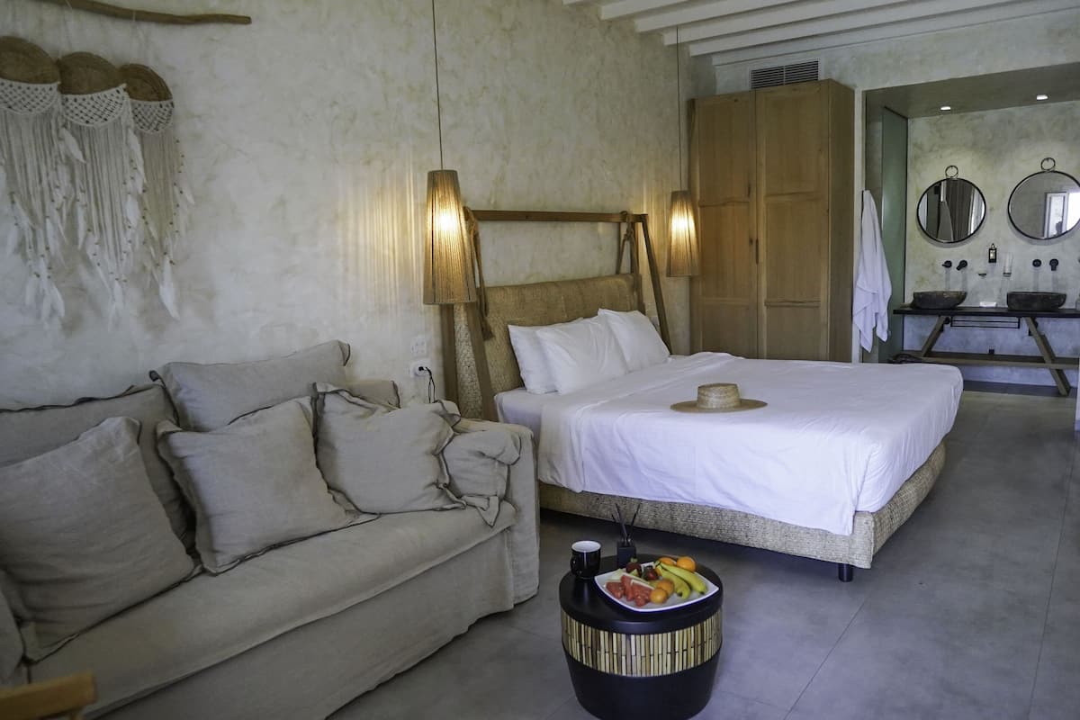 A bedroom with a large bed, a sofa, a bedside table with fruit, and a mirror above the sink.