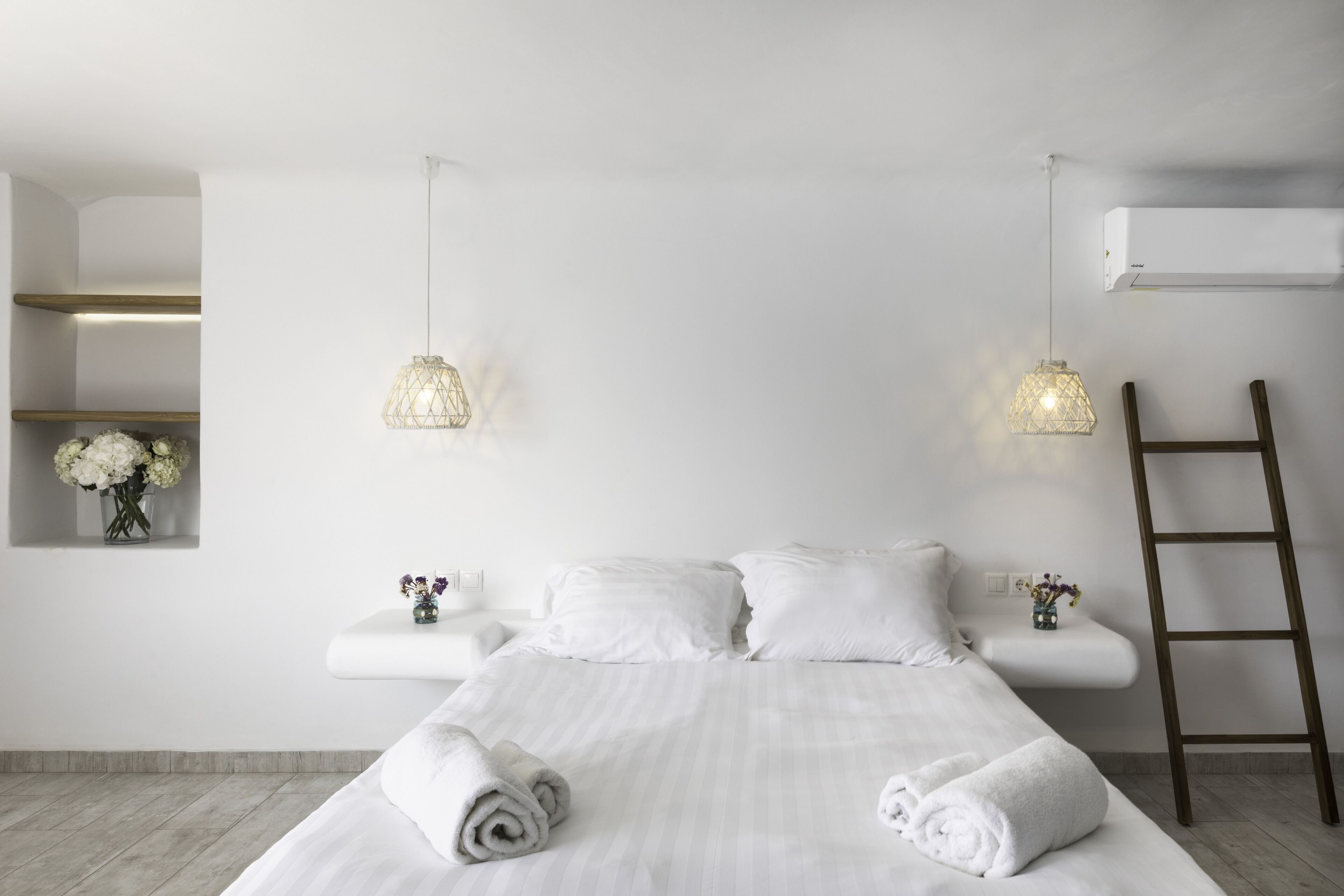 A minimalist hotel room with a large bed, white linens, two bedside tables with flower vases, and a wooden ladder.