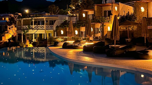 A nighttime pool area with a clear view of the pool, lounge chairs, umbrellas, and surrounding buildings. - Panormos Village Seaside Resort (Mykonos)