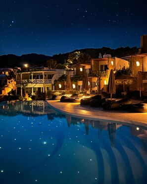A nighttime pool area with a clear view of the pool, lounge chairs, umbrellas, and surrounding buildings. - Panormos Village Seaside Resort (Mykonos)