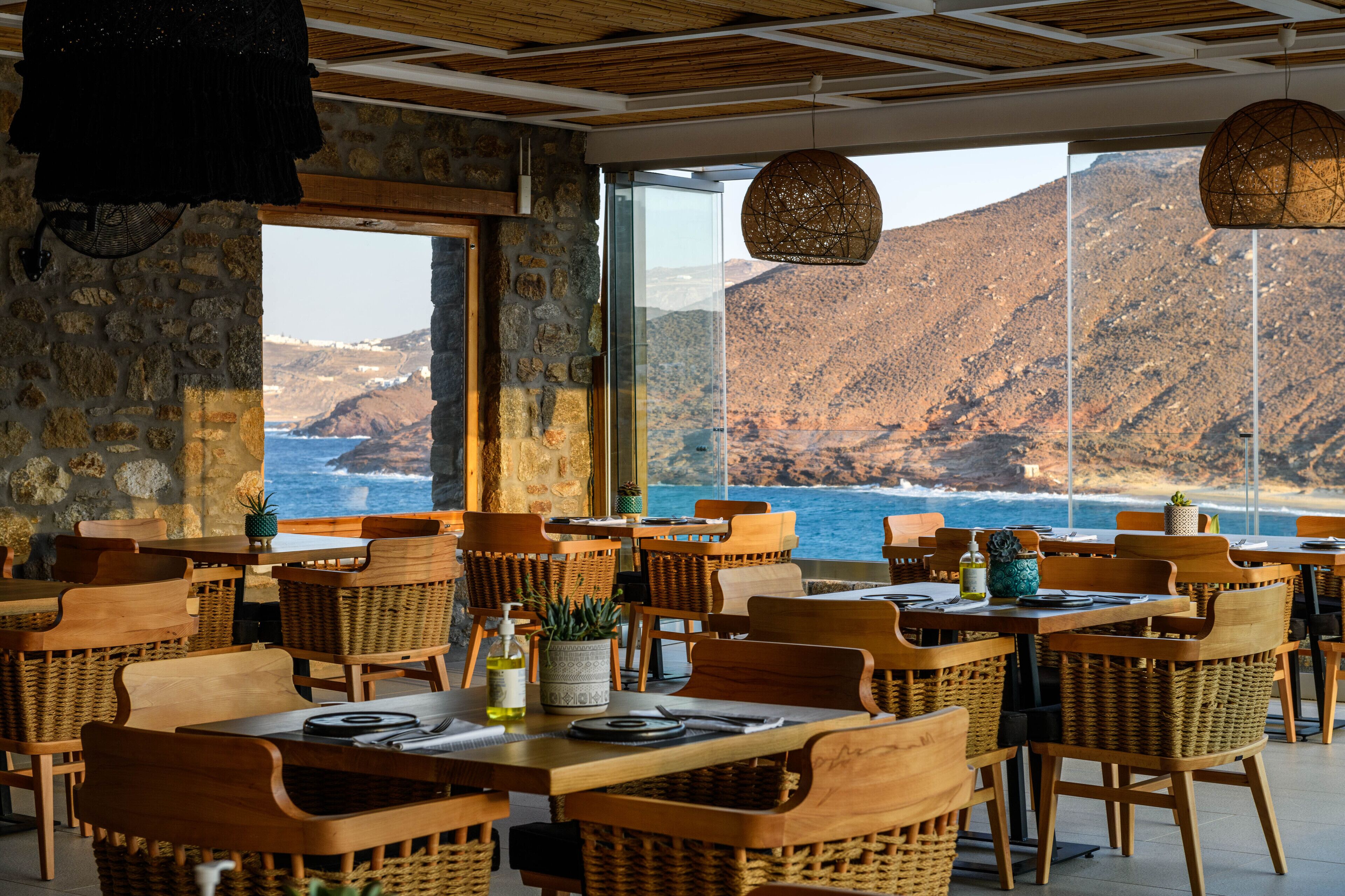 Un restaurante con mobiliario de mimbre, paredes de piedra y amplias ventanas con vistas al océano.