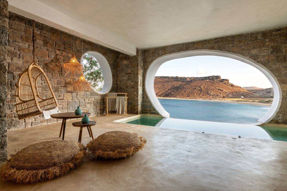 A room with a large window overlooking a body of water and a mountainous landscape. There is a hanging chair, a small round table with a bottle, and two round cushions on the floor.