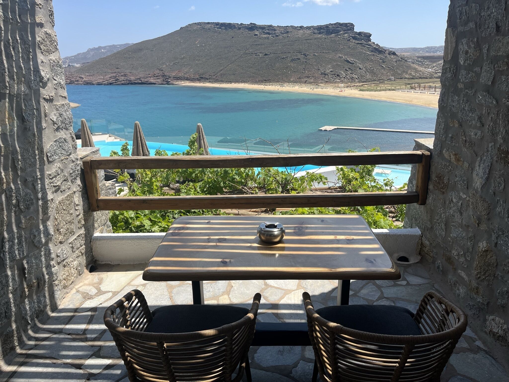 A balcony with a table and two wicker chairs, overlooking a beach and a mountain.