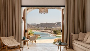 A room with a view of a beach and mountains, featuring a sofa, a chair, and a table with decorative items. - Panormos Village Seaside Resort (Mykonos)