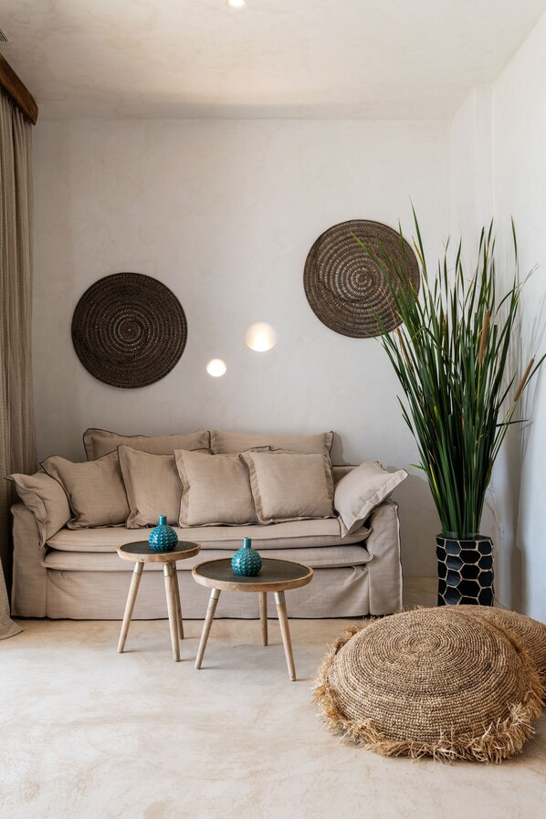 A living room with a beige sofa, two small round tables, and a large potted plant. - Panormos Village Seaside Resort (Mykonos)