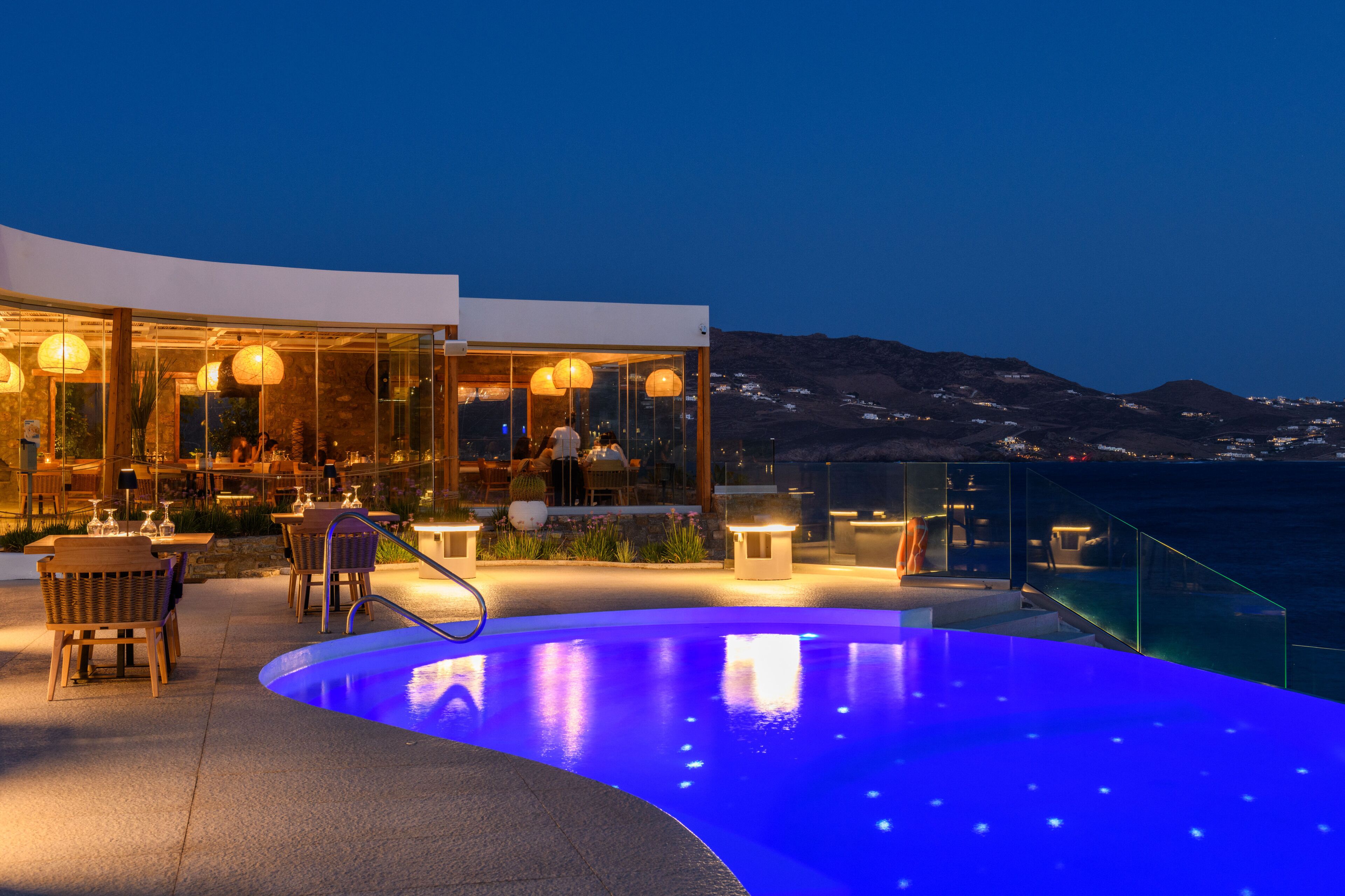 An infinity pool with a view of the sea and mountains, outdoor seating, and a restaurant with glass walls.