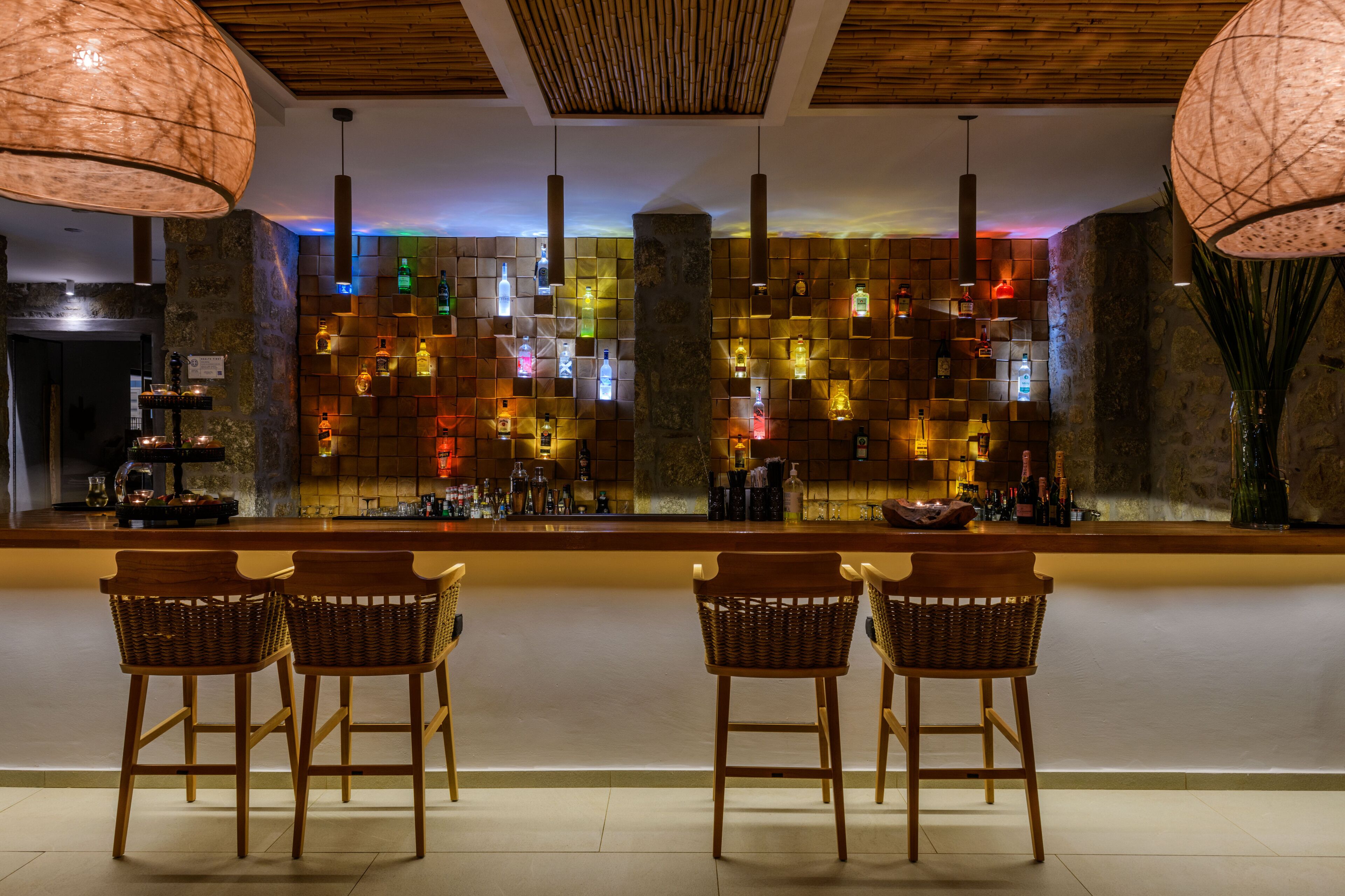 Una zona de bar con pared de piedra, barra de madera y tres sillas de mimbre.