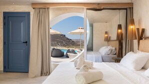 A hotel room with a bed, towels, a view of a pool and mountains, and a blue door. - Panormos Village Seaside Resort (Mykonos)
