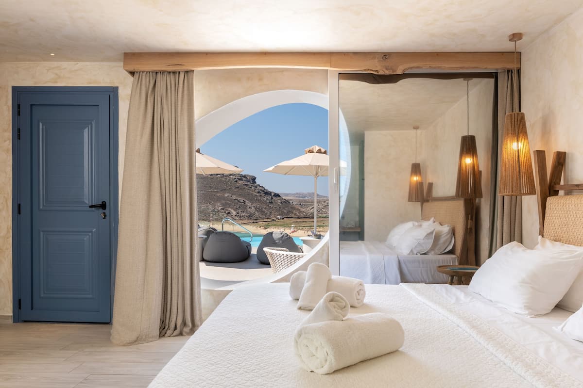 A hotel room with a bed, towels, a view of a pool and mountains, and a blue door.