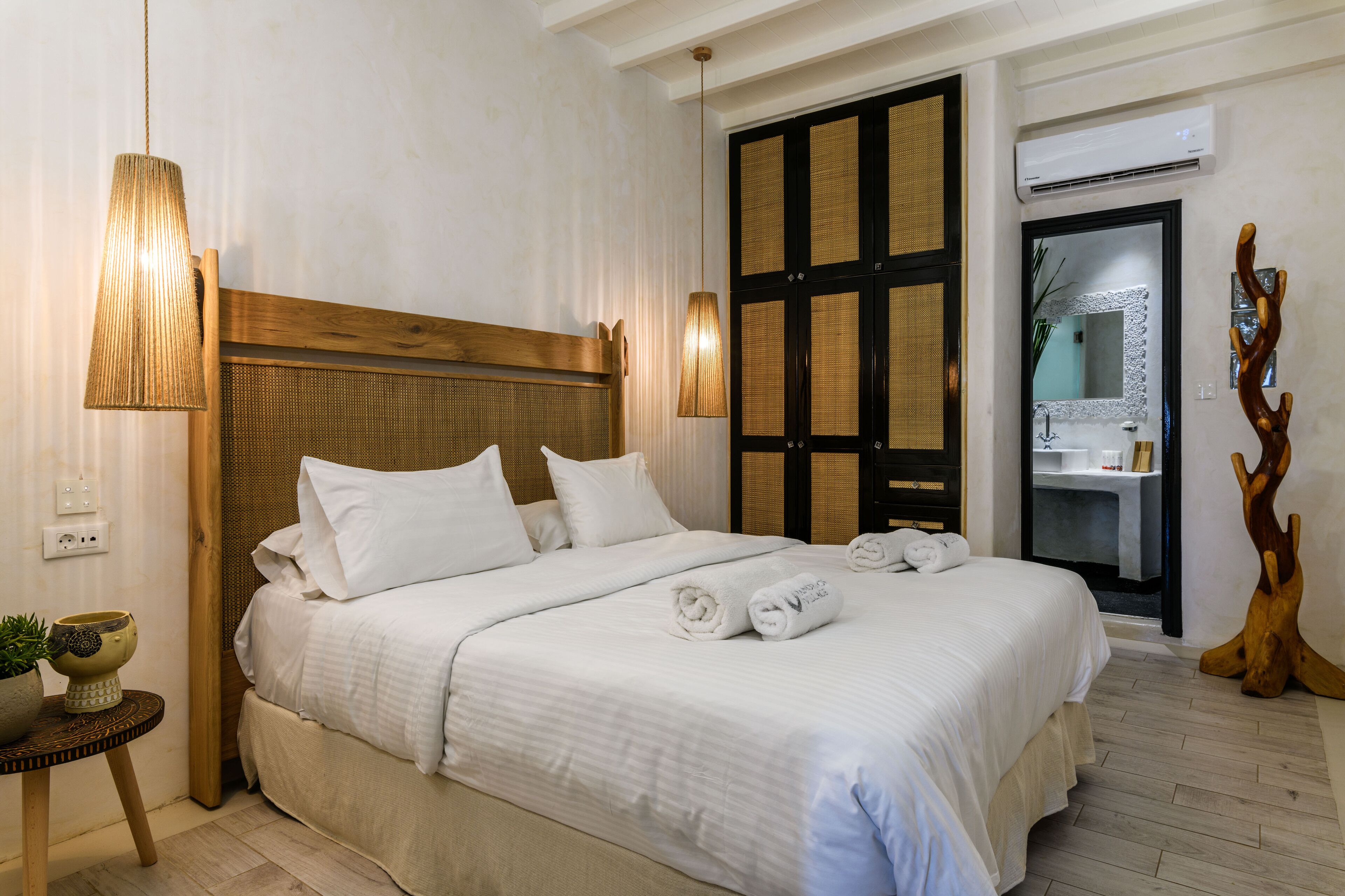 A neatly made bed with white linens and two rolled towels, a wooden headboard, a bedside lamp, and a small table with a plant.
